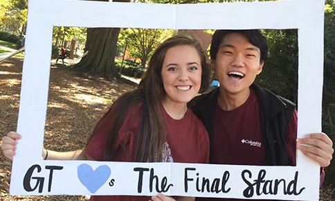 One female and one male student holding a photo frame that reads GT a heart the letter s The Final Stand.